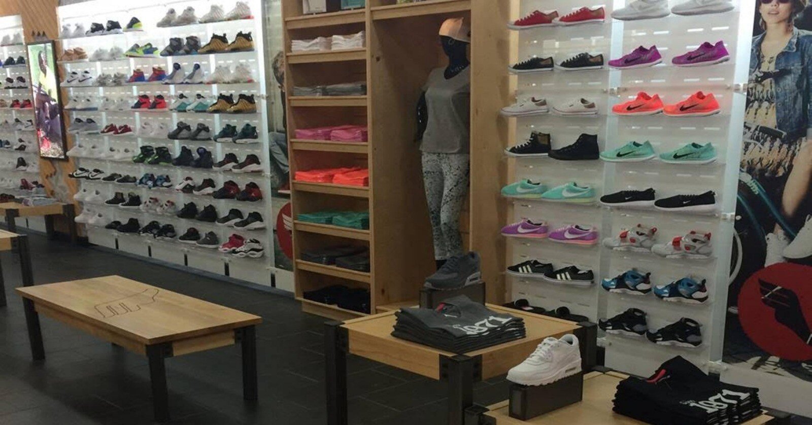 Wall of sneakers of the wall with a cubby of t-shirts and a mannequin between them. In front of the wall are tables with shirts folded on them. 